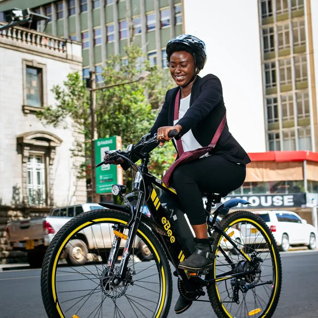 A Kenyan businesswoman in a black riding an eBee eBX through he bustling streets of Nairobi, smiling as she enjoys a quiet commute home or to the office. Gliding on her eBee e-bike, she contributes to a cleaner city and a healthier planet, one sustainable ride at a time.