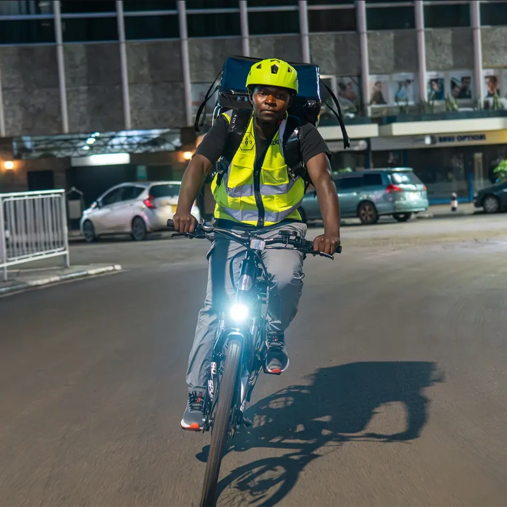 Handsome Kenyan man riding an eBee electric bicycle through the bustling streets of Nairobi on a cool evening. Headlight of the eBee on, he smiles subtly.